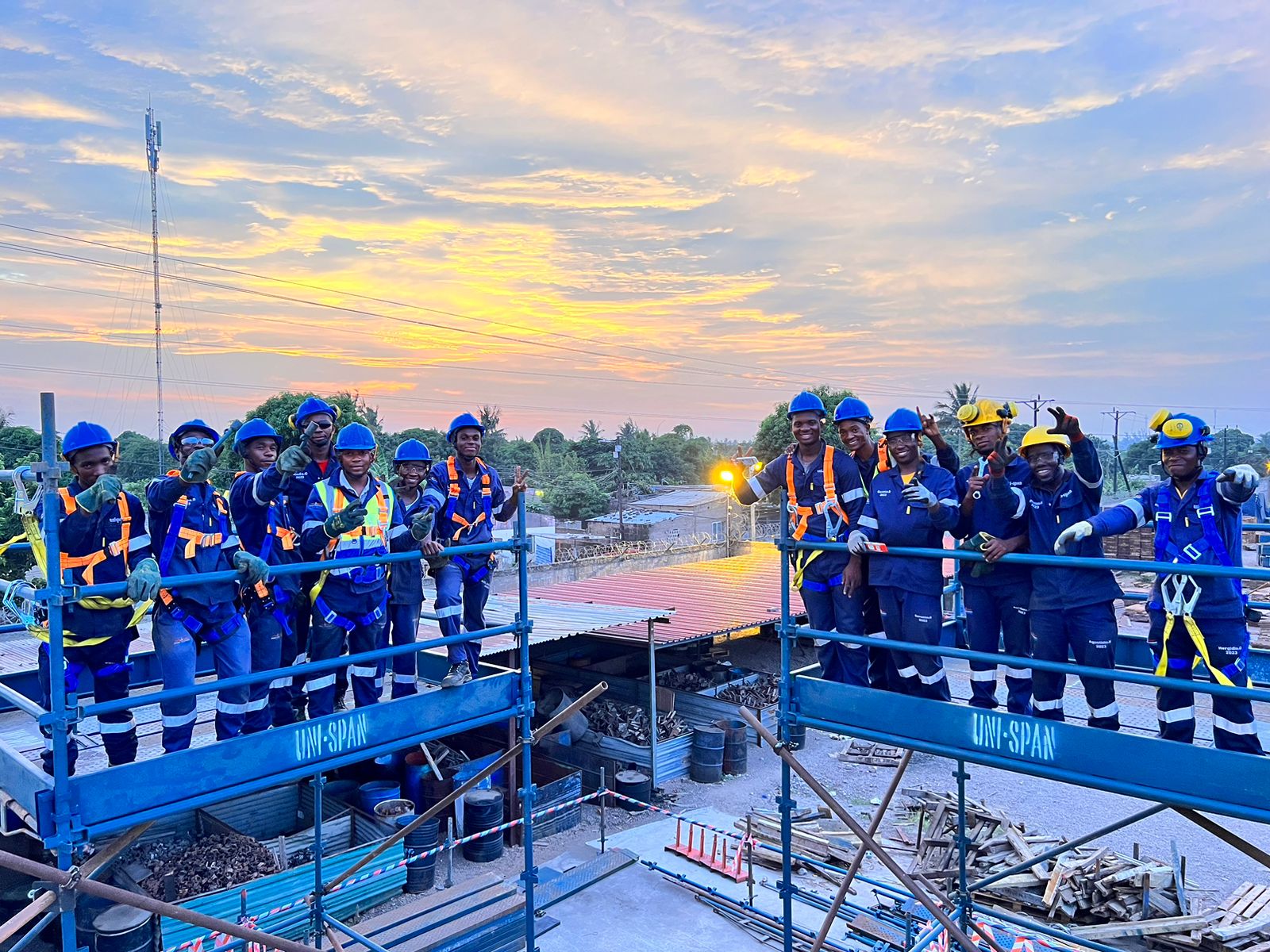 Working at Heights. A team from Uni-Span, proudly trained by TTA, stands confidently on a construction site in Mozambique. Dressed in blue uniforms and safety helmets, they showcase a sense of achievement against a backdrop of a stunning sunset. The site, scattered with equipment and scaffolding, highlights the ongoing development and the collaboration between TTA and Uni-Span.